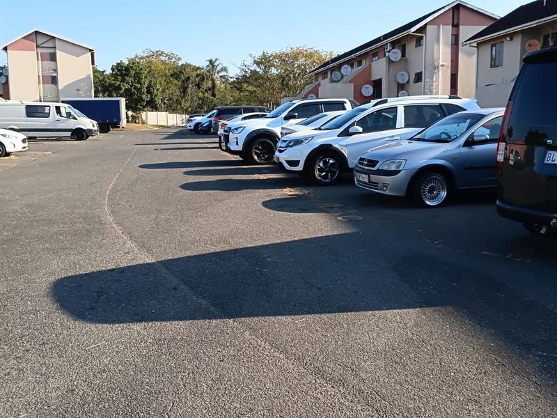 An outdoor parking area situated between multi-story apartment buildings, featuring paved surfaces and designated vehicle parking spaces.