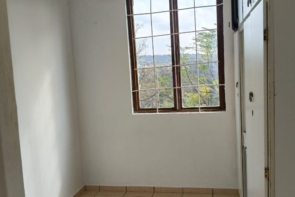 A compact, empty bedroom featuring tiled flooring, built-in cupboards, and a window with security bars offering a natural view.