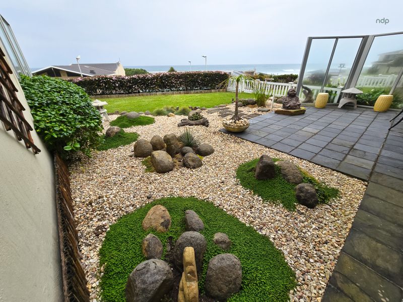 A well-maintained landscaped garden featuring a paved patio area, rock garden elements, and a glass balustrade with ocean views.