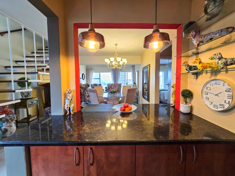 Open plan kitchen area featuring a dark granite breakfast bar, wooden cabinetry, and pendant lighting, looking through to the dining and living space.