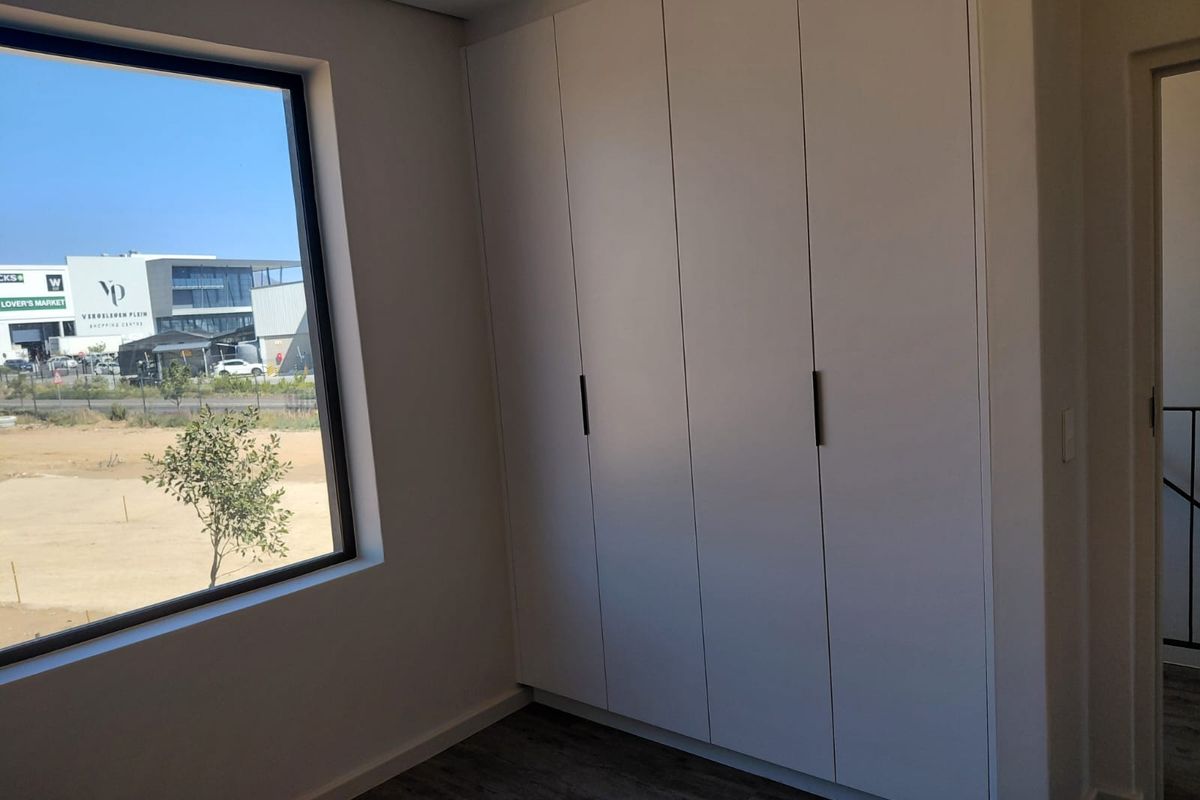A bright, modern bedroom featuring sleek built-in cupboards, high-quality laminate flooring, and a large window offering natural light and an exterior view.