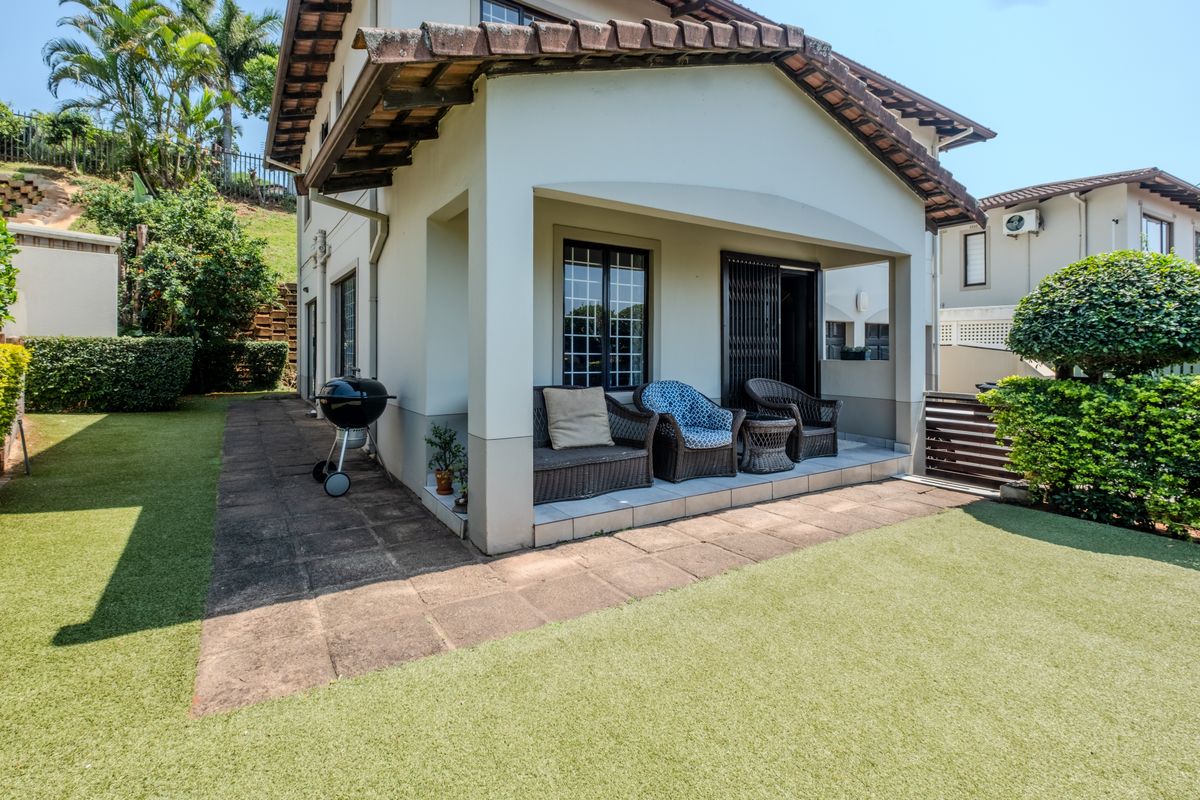 Rear view of the townhouse featuring a covered patio, paved walkway, and a well-maintained artificial lawn area.