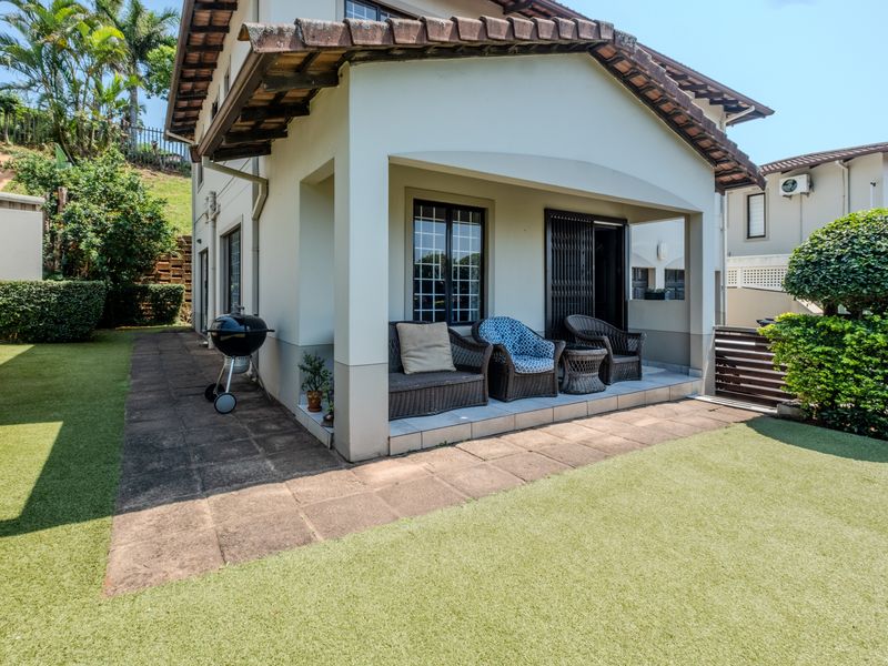 Rear view of the townhouse featuring a covered patio, paved walkway, and a well-maintained artificial lawn area.