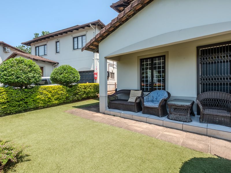 A covered patio area featuring tiled flooring and a security gate, overlooking a well-maintained garden with artificial lawn.
