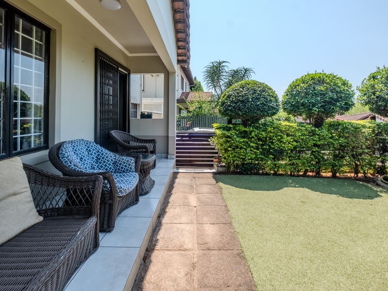 A covered patio area featuring tiled flooring and sliding door access, leading out to a private garden with artificial lawn and established hedges.