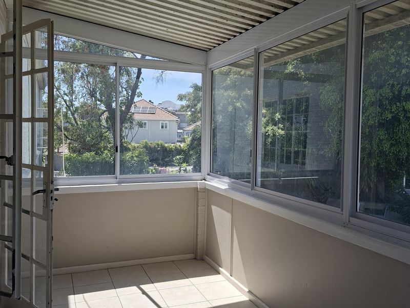A bright, enclosed balcony or sunroom featuring tiled flooring, large sliding glass windows, and a corrugated ceiling, offering a versatile outdoor living space.