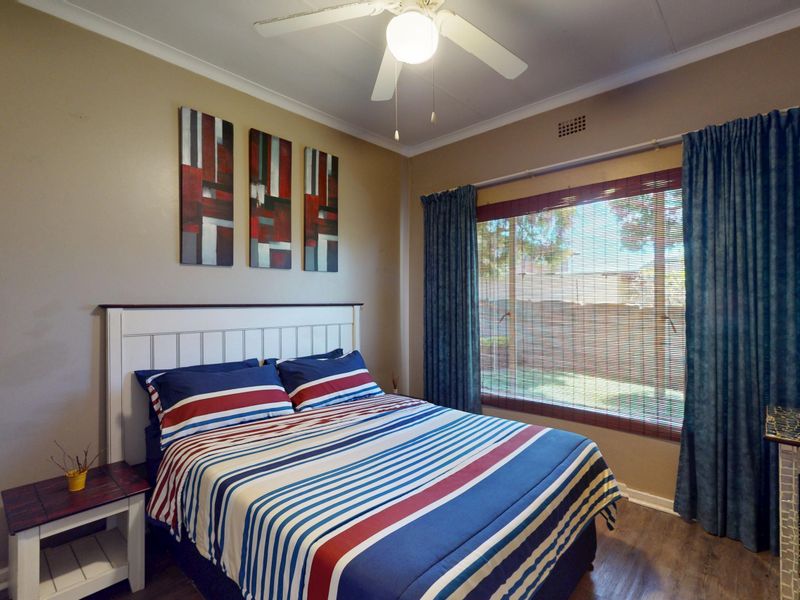A comfortable bedroom featuring laminate flooring, a ceiling fan, and large windows with blinds and curtains, offering a functional and well-lit living space.