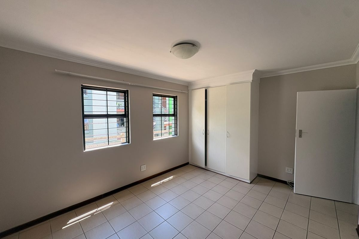 A bright, empty bedroom featuring neutral walls, tiled flooring, and built-in cupboards with security bars on the windows.