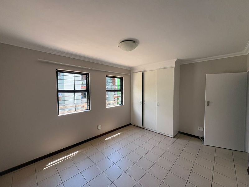 A bright, empty bedroom featuring neutral walls, tiled flooring, and built-in cupboards with security bars on the windows.