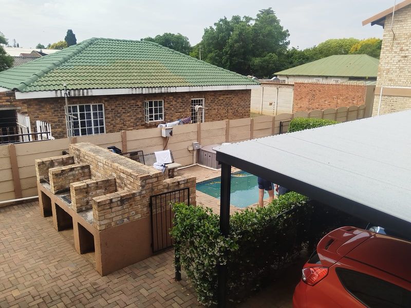 Outdoor entertainment area featuring a built-in brick braai, swimming pool, and covered carport parking space within a paved courtyard setting.