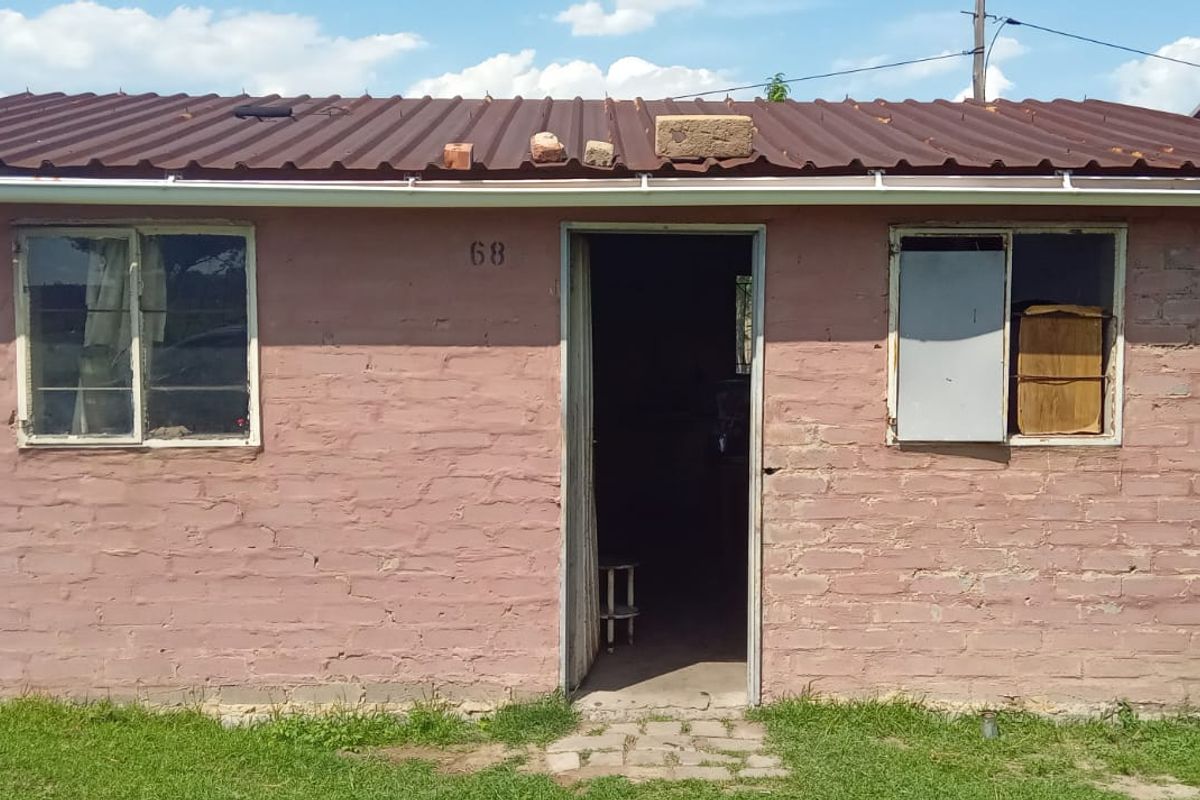 Front elevation of a single-story brick house featuring a corrugated metal roof and basic window frames, showing signs of significant wear and maintenance requirements.