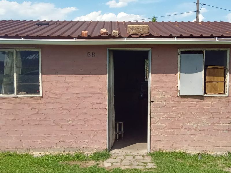 Front elevation of a single-story brick house featuring a corrugated metal roof and basic window frames, showing signs of significant wear and maintenance requirements.