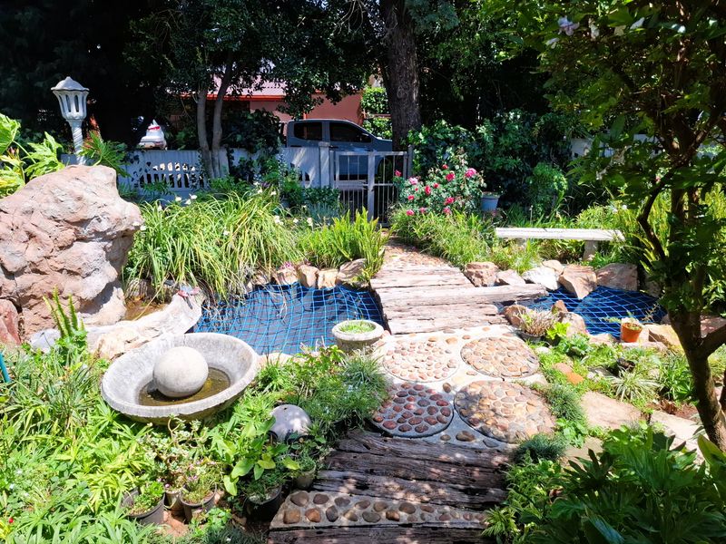 A lush garden featuring a pond with a wooden bridge, stone pathways, and a decorative water feature, enclosed by a perimeter wall and gate.