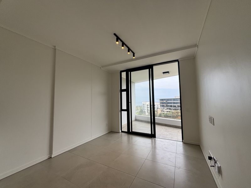 A bright, empty bedroom featuring modern tiled flooring, track lighting, and a sliding glass door providing direct access to a private balcony.