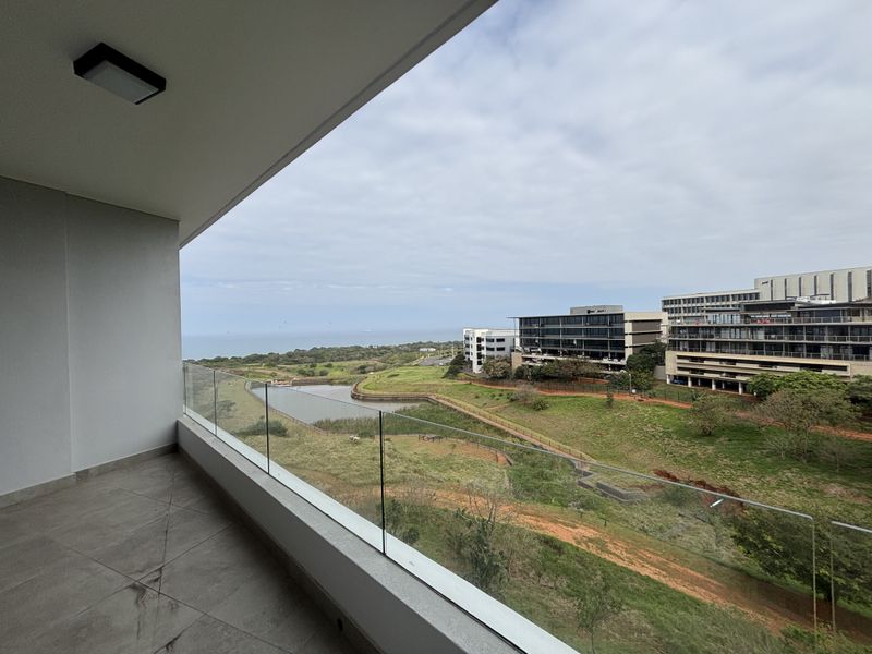A modern covered balcony featuring tiled flooring and a frameless glass balustrade, offering expansive views over the surrounding landscape and nearby buildings.