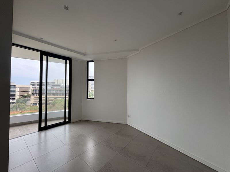 A bright, modern, and empty living area featuring large windows, sliding glass doors leading to a balcony, and neutral tiled flooring.
