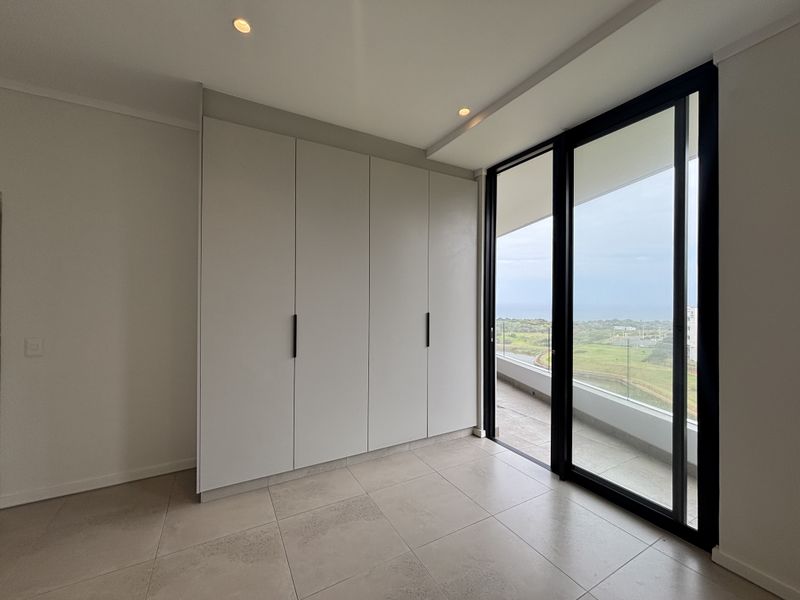 A bright, modern bedroom featuring sleek built-in cupboards, large sliding glass doors leading to a balcony, and neutral tiled flooring with scenic views.