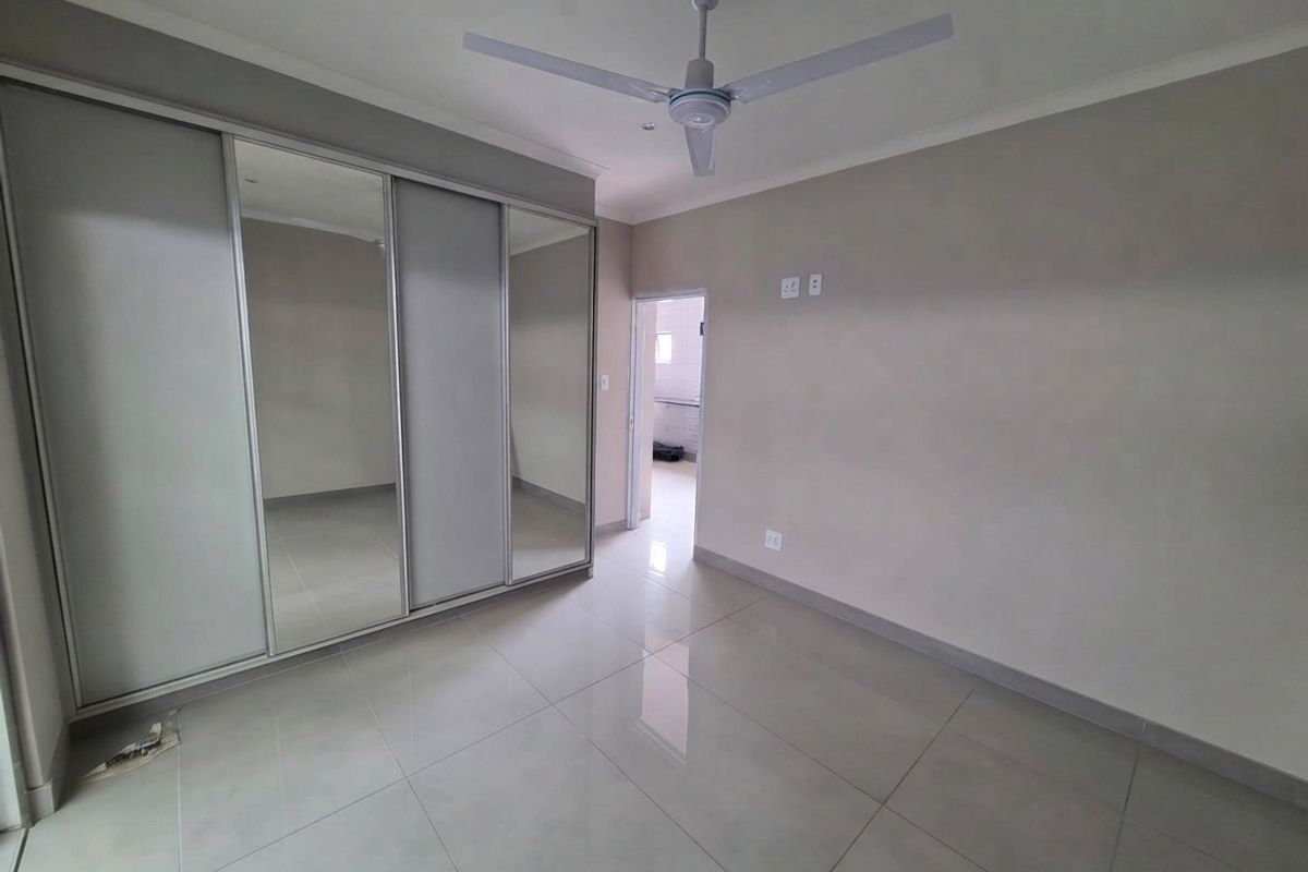 A spacious, empty bedroom featuring neutral tiled flooring, a large built-in mirrored sliding wardrobe, and a ceiling fan.