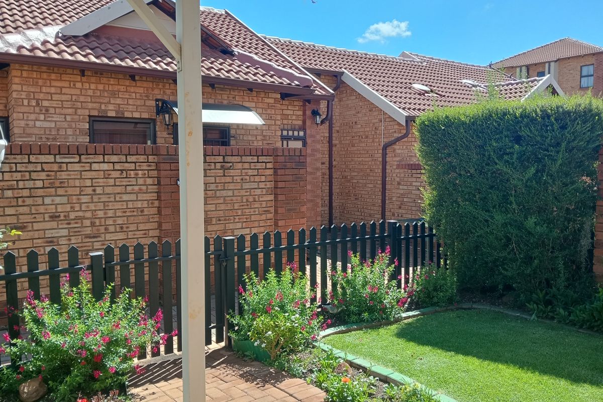 A well-maintained rear exterior view featuring a face brick facade, tiled roof, paved patio area, and a neat garden with a picket fence.