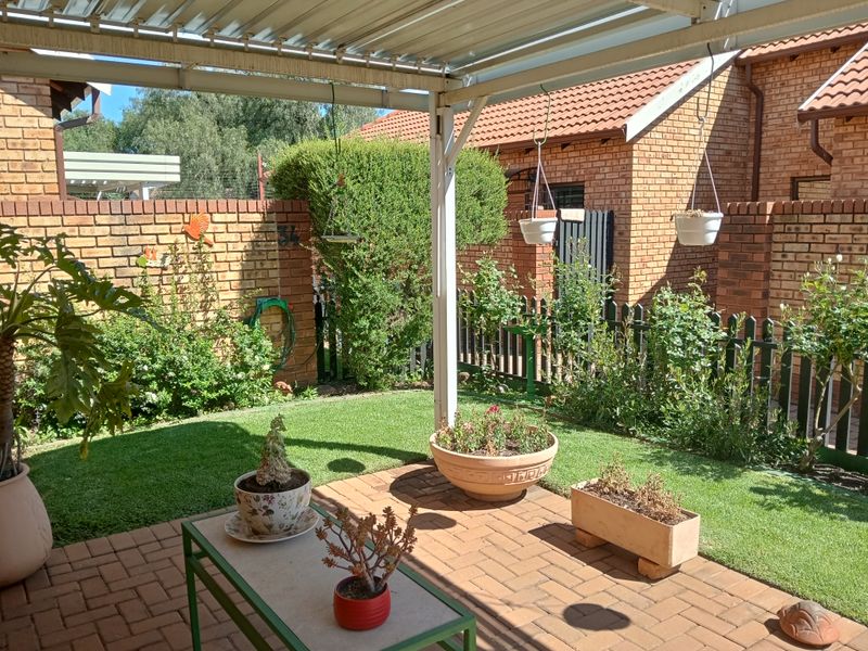 A well-maintained covered patio area featuring brick paving and a metal roof structure, overlooking a private garden with brick boundary walls.