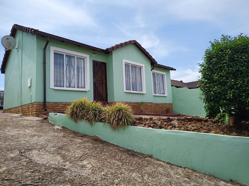 Front elevation of a single-story house featuring a green painted facade, brick plinth, tiled roof, and a concrete driveway with a landscaped retaining wall.