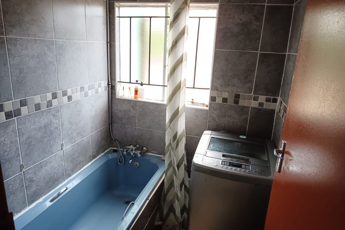 A functional bathroom featuring grey wall tiles and a blue bathtub, with a window providing natural light and space for a washing machine.