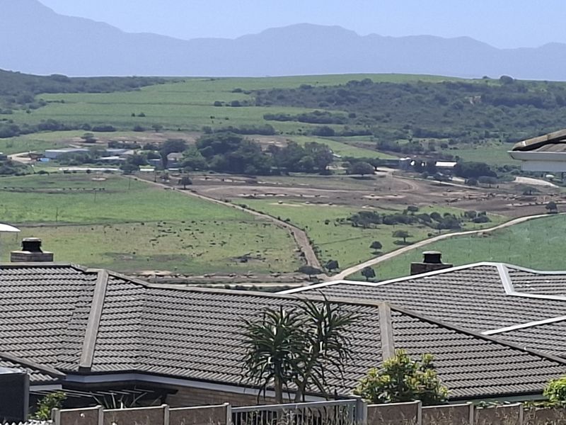 Elevated view overlooking a rural landscape with rolling green hills and distant mountains, featuring residential tiled roof structures in the foreground.