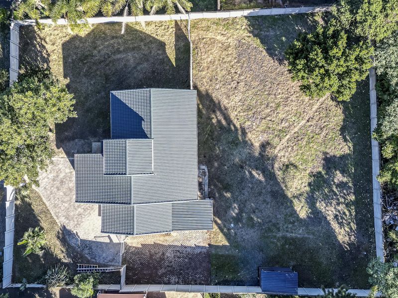 Aerial view of the property showing the house footprint, metal roof, paved driveway, and surrounding garden area within the fenced perimeter.