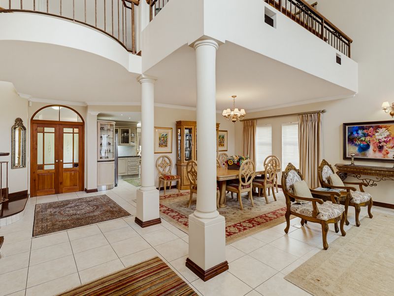 Spacious open-plan living and dining area featuring a double-height ceiling, elegant columns, a grand staircase, and a mezzanine landing overlooking the main floor.