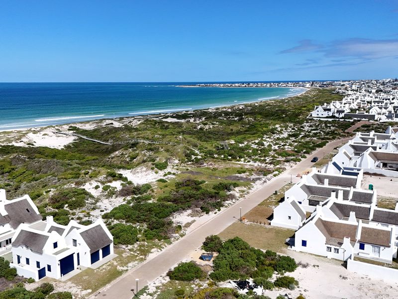 An aerial view of a coastal residential estate featuring white houses with thatched roofs, situated near the beach and natural dunes.