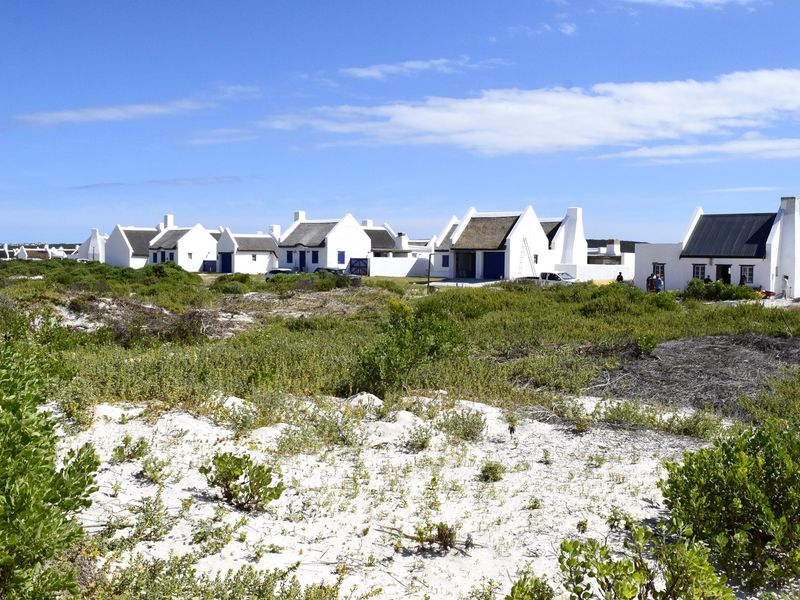 A view of a coastal residential development featuring traditional white cottages with thatched roofs set within a natural sandy landscape.