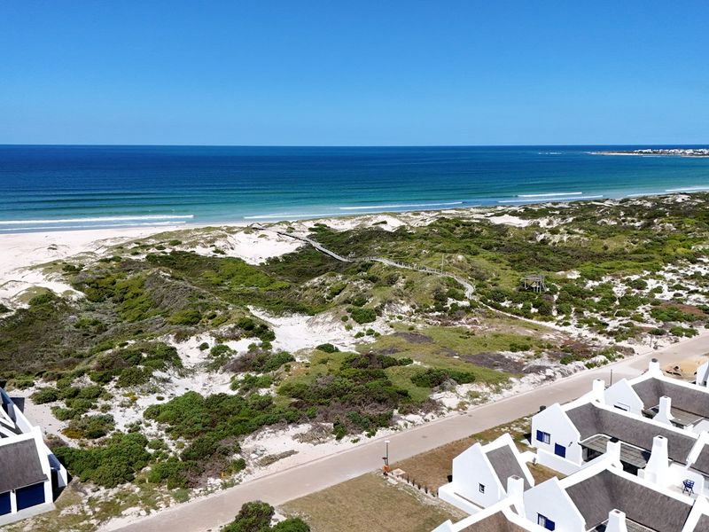 Aerial view of a coastal residential development featuring white houses with thatched roofs, adjacent to natural dunes, vegetation, and a sandy beach.