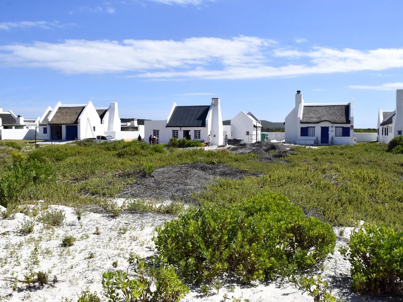 A view of a residential development featuring multiple white cottages with thatched roofs and blue shutters, set within a natural coastal landscape.