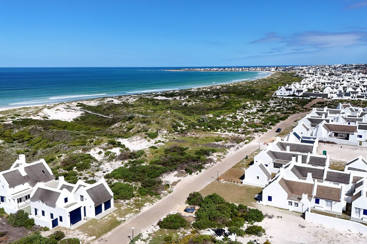 An aerial view of a coastal residential estate featuring white houses with thatched roofs, situated near the beach and natural dunes.