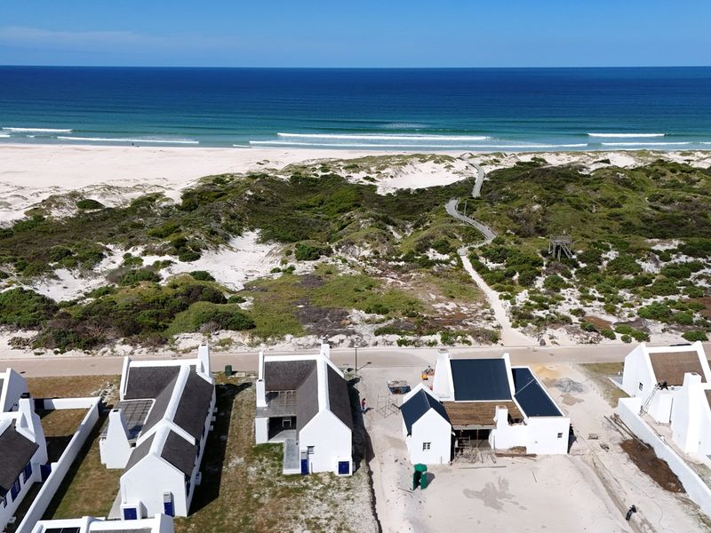 Aerial view of a coastal residential development featuring white buildings with thatched roofs, situated near sand dunes and the ocean.