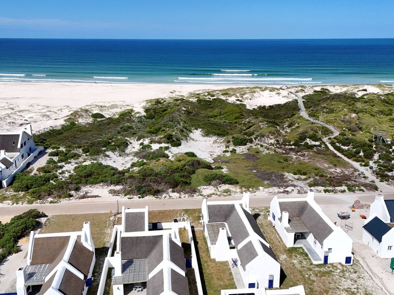 Aerial view of a coastal residential development featuring white buildings with thatched roofs, situated directly adjacent to the beach and dunes.