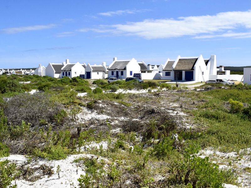 A view of a residential estate featuring white cottages with traditional thatched roofs, set within a natural coastal landscape with sandy terrain and native vegetation.