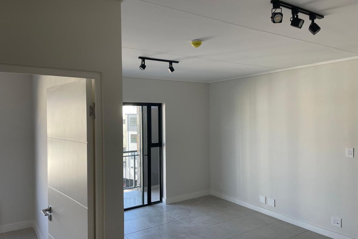 A bright, empty living area featuring modern track lighting, neutral tiled flooring, and a sliding glass door leading to a balcony.