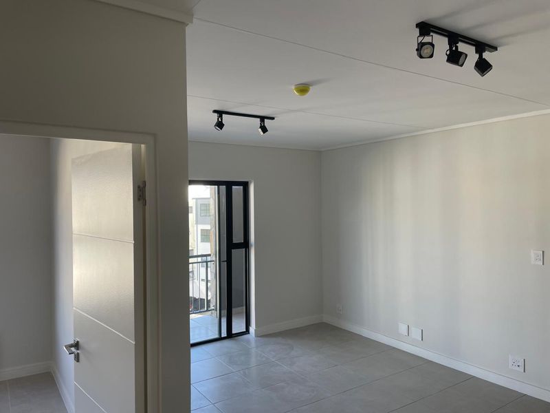 A bright, empty living area featuring modern track lighting, neutral tiled flooring, and a sliding glass door leading to a balcony.