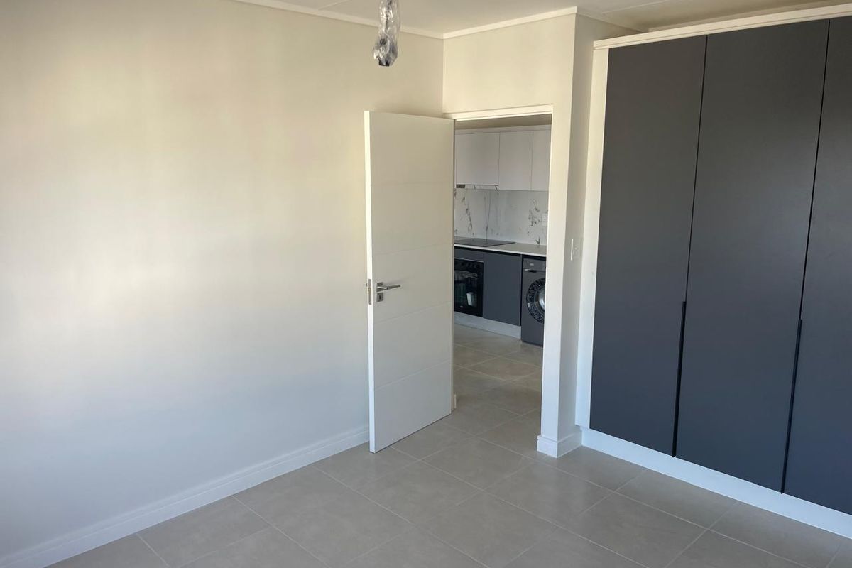 A clean, empty bedroom featuring modern grey built-in cupboards, neutral tiled flooring, and a view through to the adjacent kitchen area.