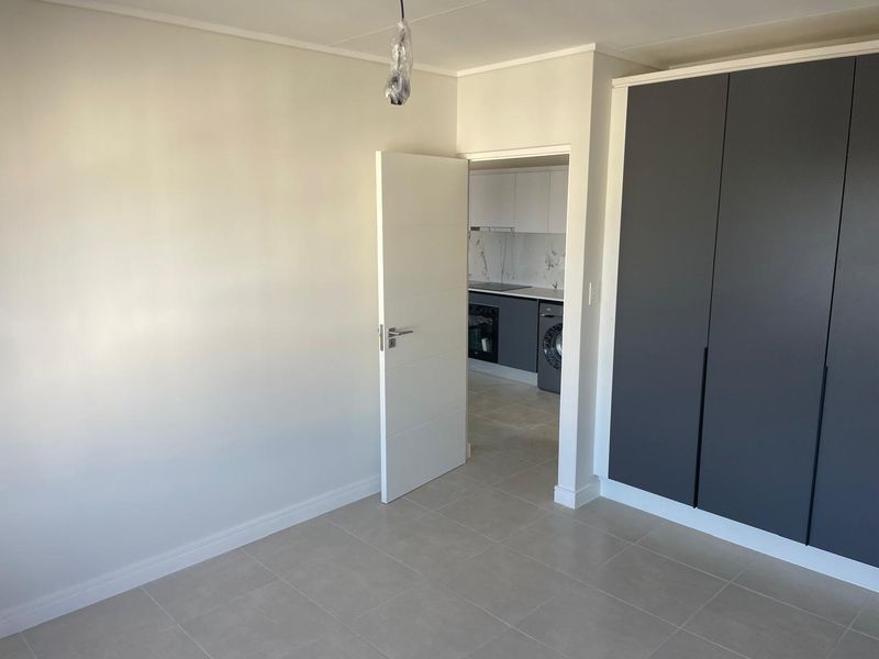 A clean, empty bedroom featuring modern grey built-in cupboards, neutral tiled flooring, and a view through to the adjacent kitchen area.