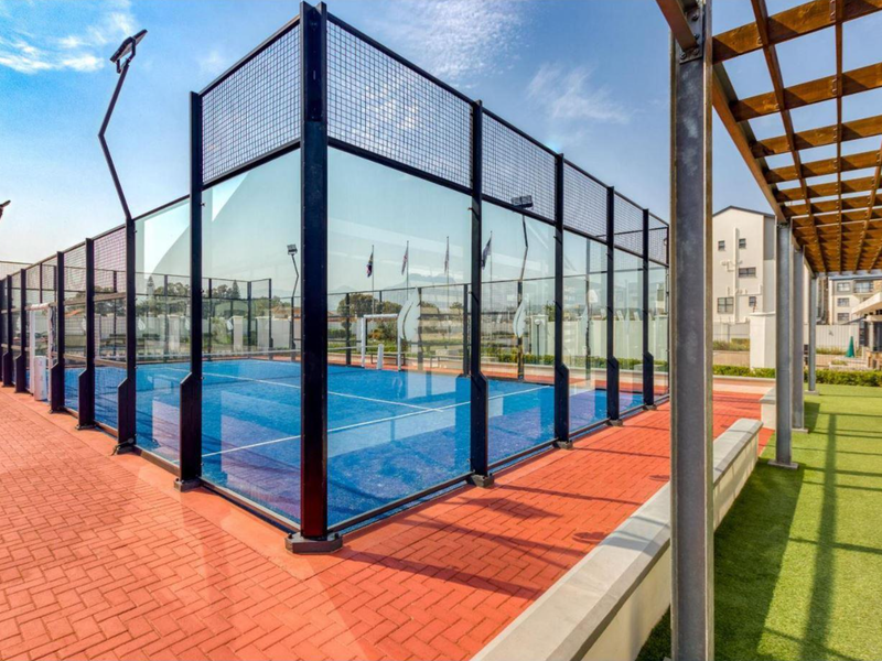 A well-maintained outdoor padel court featuring glass perimeter fencing, floodlights, and adjacent paved and artificial turf areas with a timber pergola.