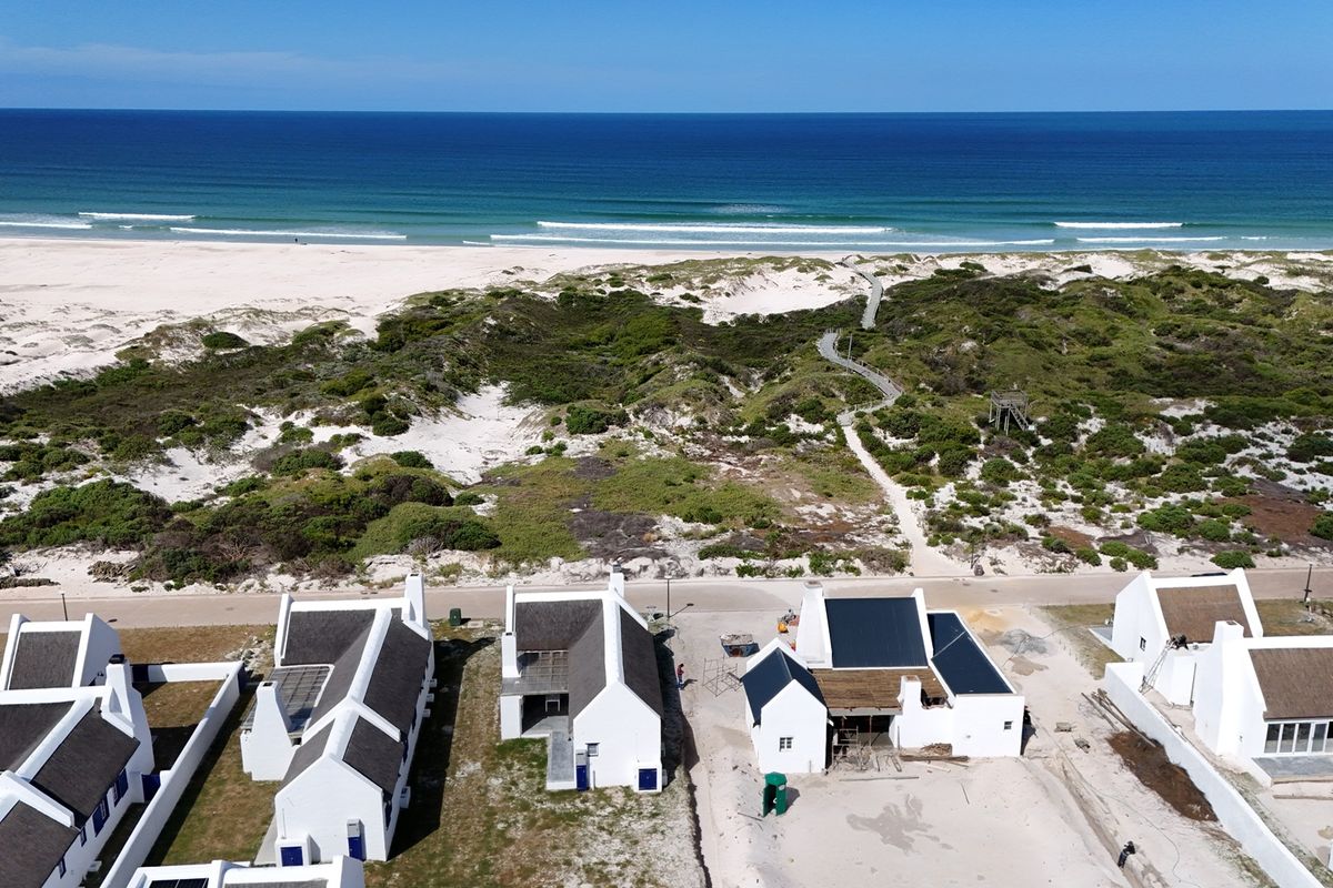 Aerial view of a coastal residential development featuring white buildings with thatched roofs, situated near sand dunes and the ocean.