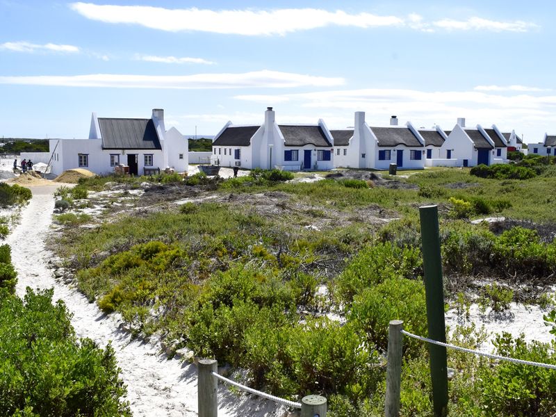 A view of a coastal residential development featuring multiple white cottages with thatched roofs and blue accents, set within a natural sandy landscape.