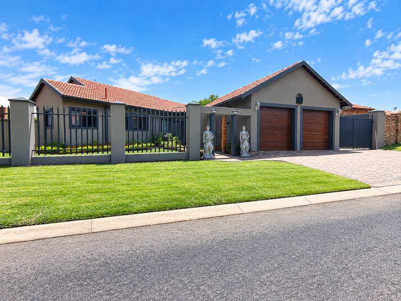 A well-maintained front elevation featuring a double garage, paved driveway, manicured lawn, and secure metal perimeter fencing with brick pillars.