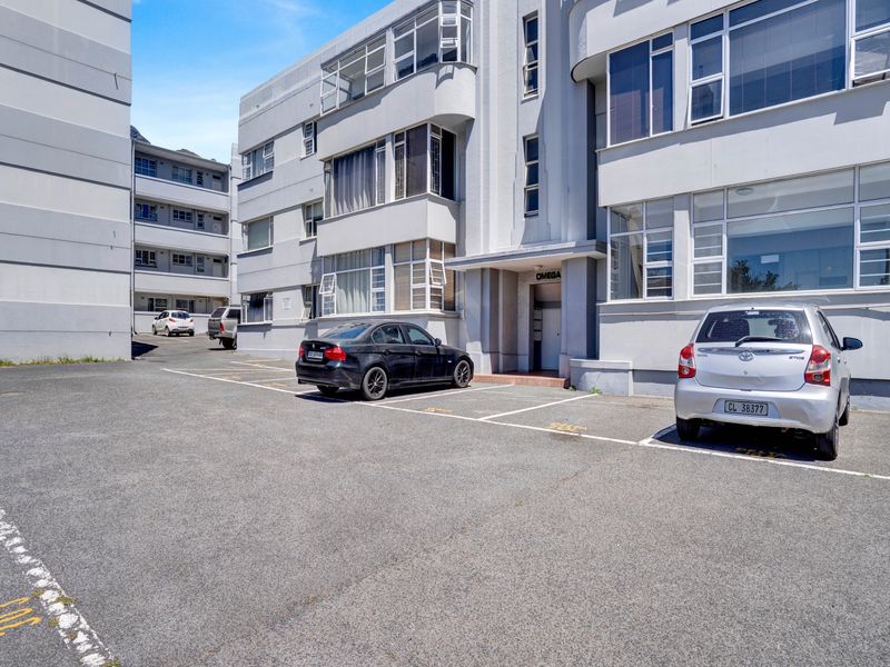 Exterior view of the apartment building entrance featuring a paved parking area and multi-story facade with large windows.
