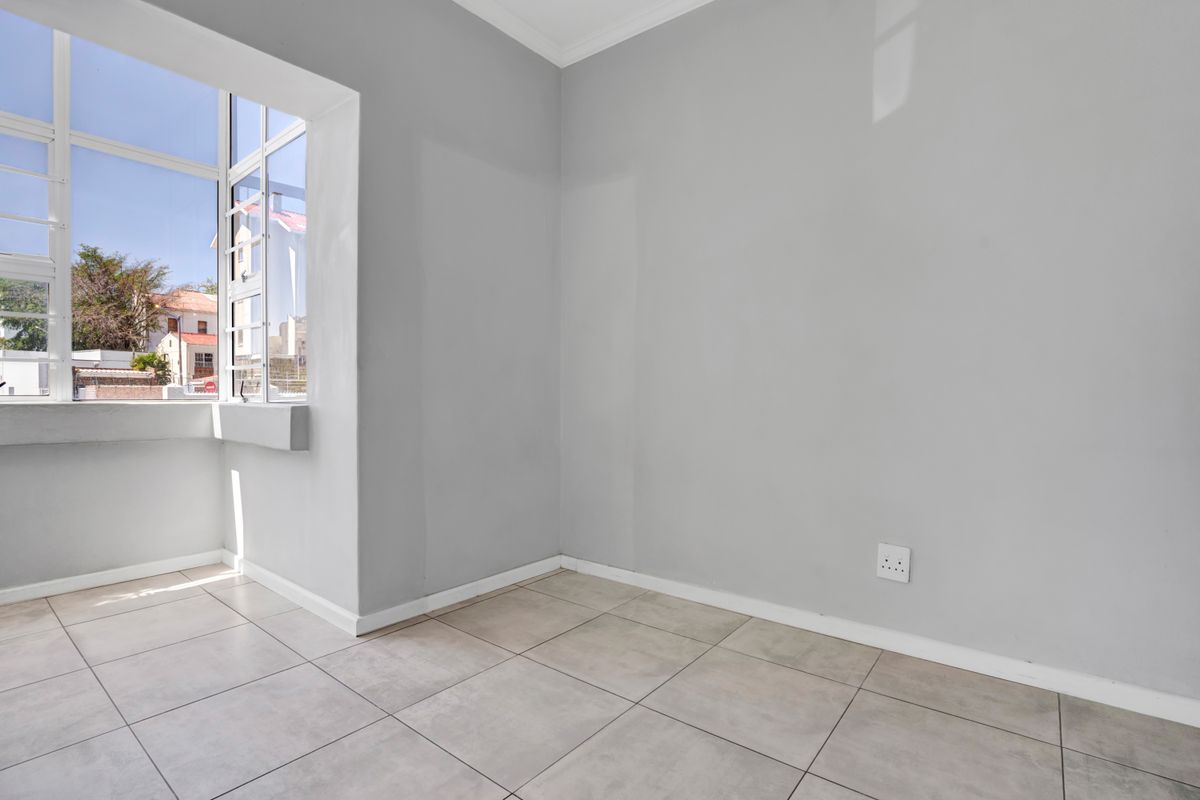 A bright and empty bedroom featuring neutral grey walls, large windows allowing natural light, and clean tiled flooring.