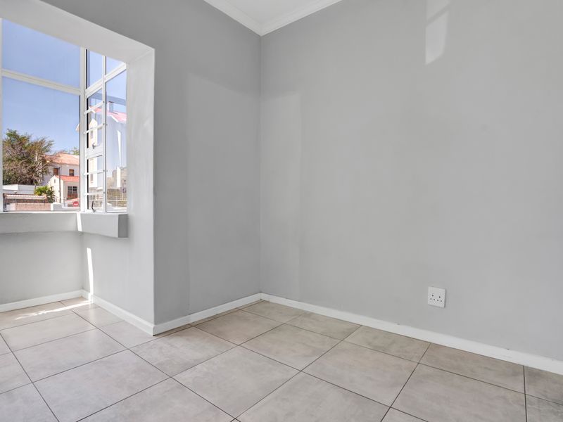 A bright and empty bedroom featuring neutral grey walls, large windows allowing natural light, and clean tiled flooring.