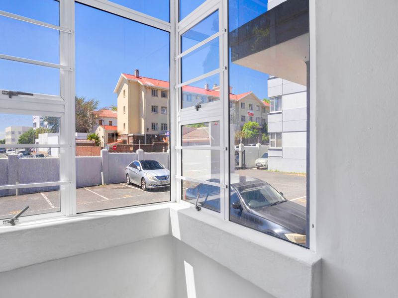 A bright, enclosed balcony area featuring large windows with security bars, offering an elevated view of the surrounding apartment complex and parking area.