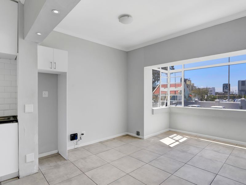 A bright, open-plan living and kitchen area featuring neutral tiled flooring, built-in cabinetry, and a large bay window with urban views.